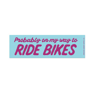 A rectangular bumper sticker with a light blue background and pink text that reads 'Probably on my way to RIDE BIKES'. The words 'RIDE BIKES' are in a larger, bold uppercase font, while the phrase 'Probably on my way to' is in a smaller script font.